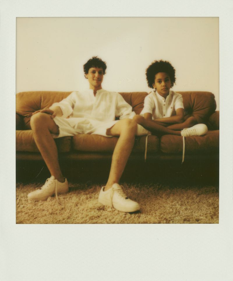 Two people sit on a brown couch in a brightly lit interior. A boy and a young man wear white shirts, shorts, and sneakers in a retro style.