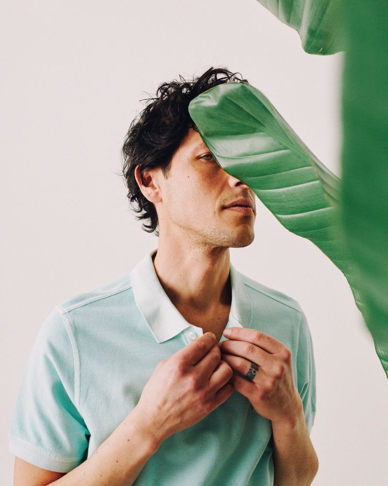 A man in a teal polo shirt is partially obscured by a large green leaf, posing against a white background in a studio portrait.