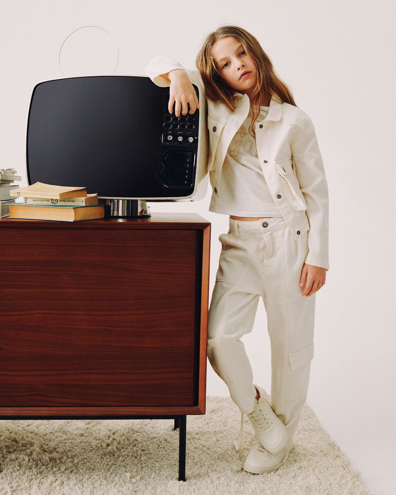 Young girl in fashion wear poses next to a retro television on a mid-century modern cabinet with books in a studio setting.
