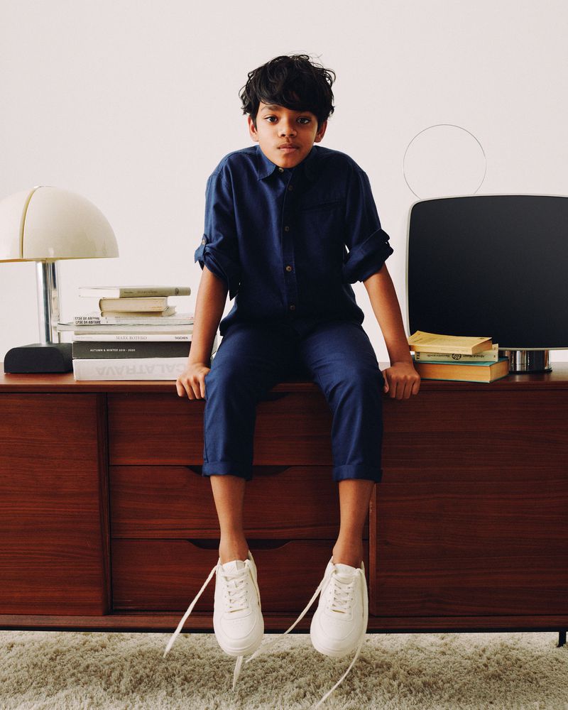 Young boy in blue outfit sits on wooden dresser with lamp, books, and television in a brightly lit room, wearing white sneakers.