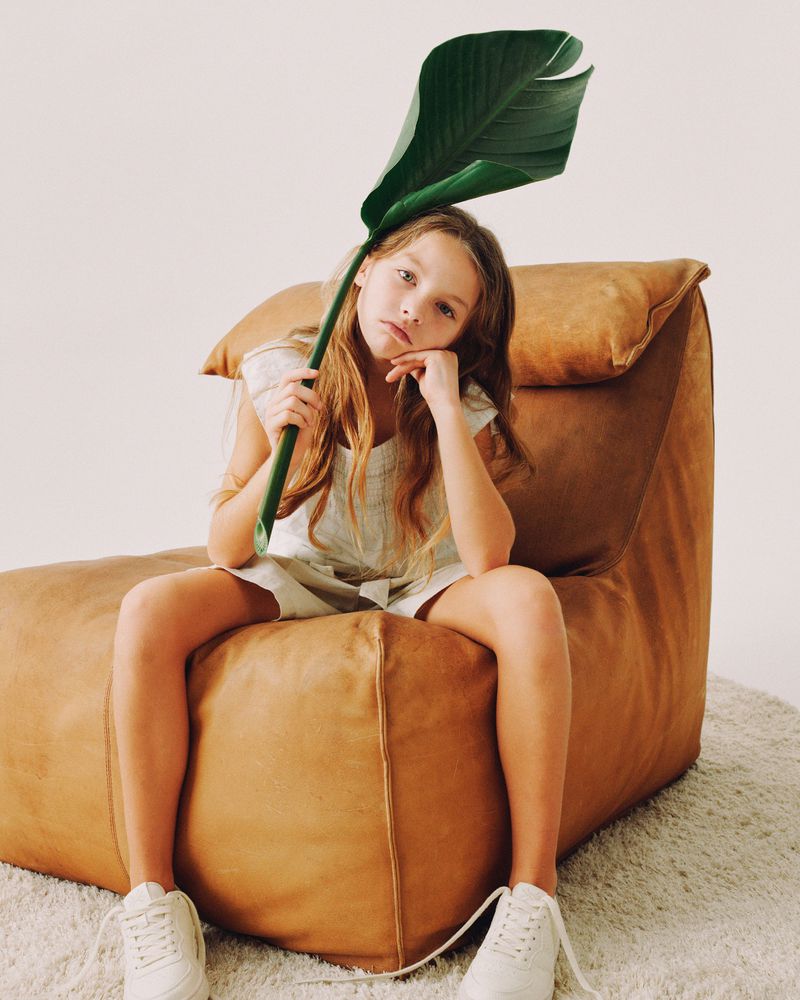 A young girl sits on a brown leather beanbag chair holding a large green leaf near her head in an indoor studio setting.