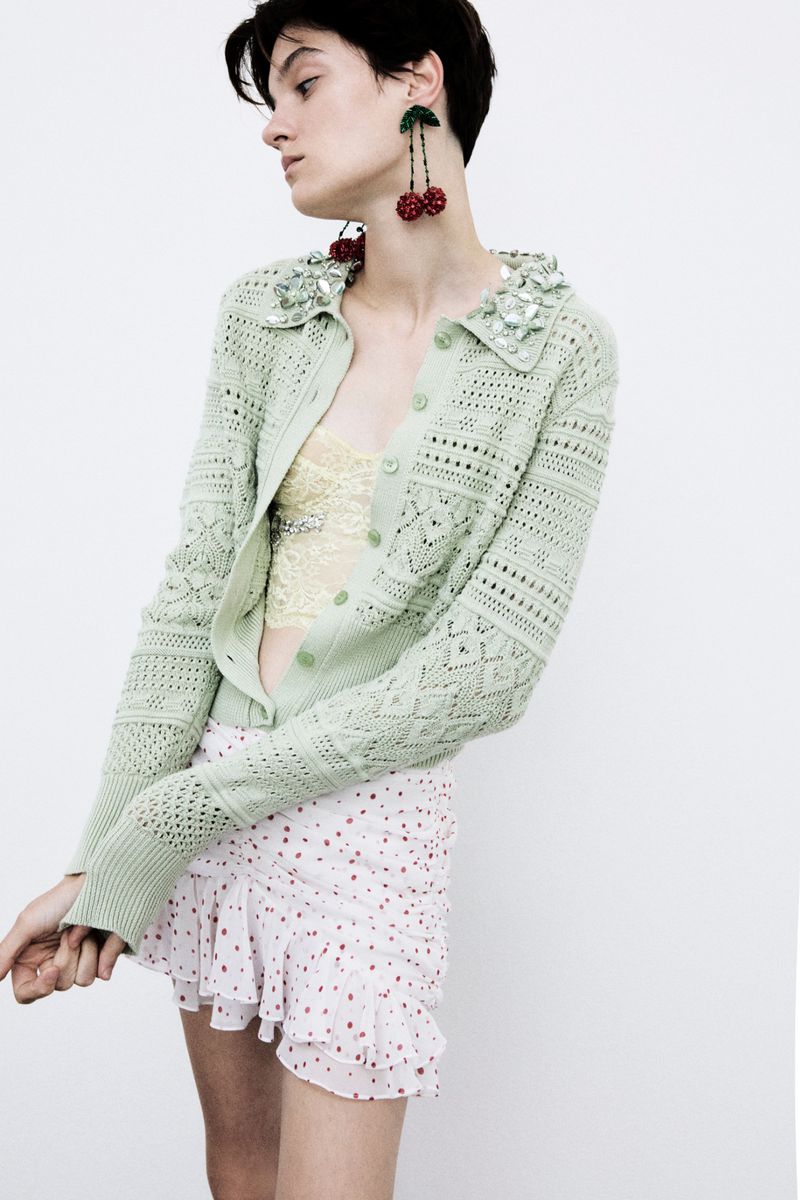 A young woman in a light green cardigan and white skirt poses in front of a white background, accessorized with cherry earrings.