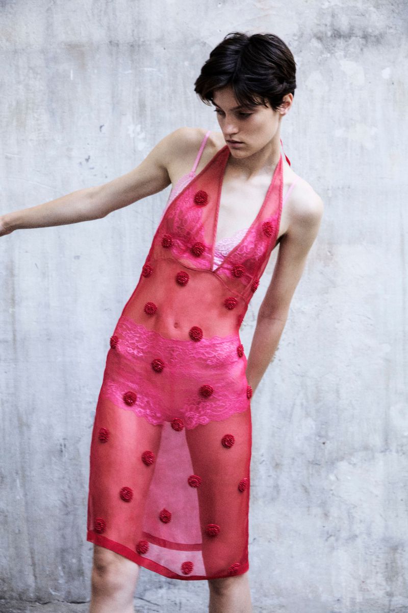 A slender woman poses in a red sheer dress adorned with roses, under soft light against a textured grey wall, fashion portrait.