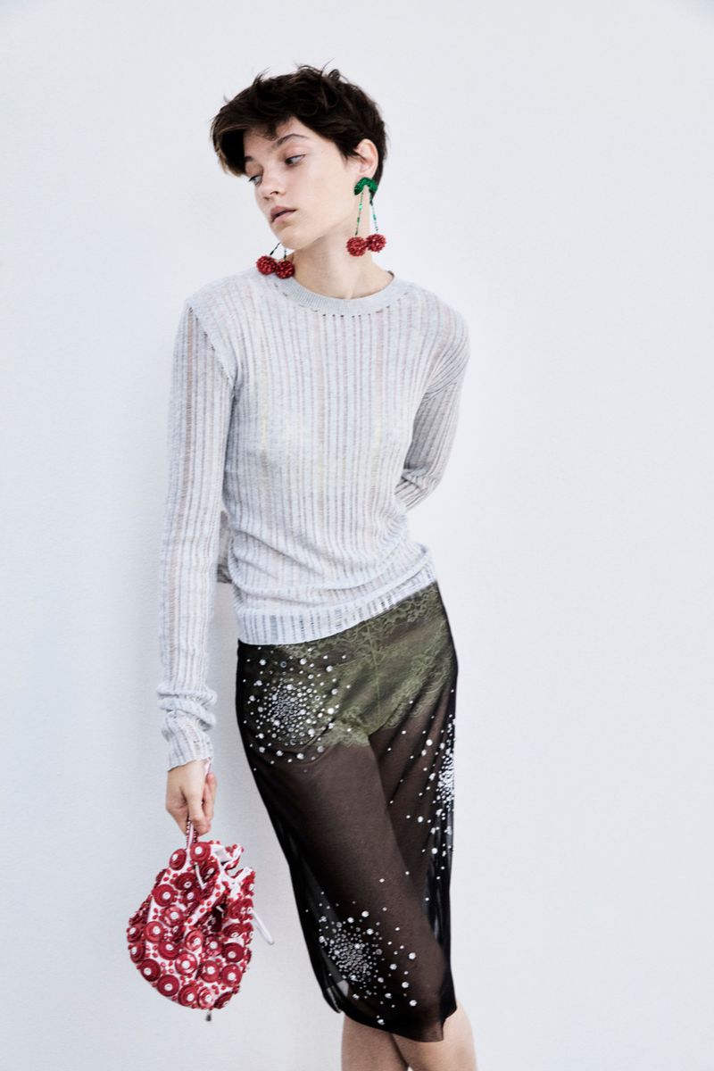 Fashion model poses against a white wall wearing a gray sweater, sparkling black skirt, cherry earrings, and holding a red bag.