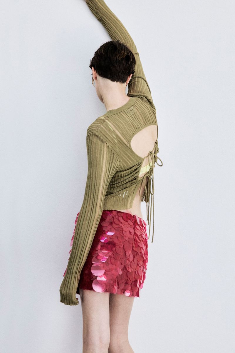 A person poses against a white wall, wearing an olive green top with a back cutout, paired with a red sequin mini skirt in a studio shot.