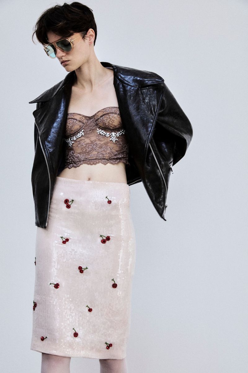 A woman in a black leather jacket, embellished lace crop top, and cherry print skirt poses against a plain background.