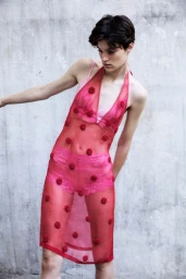 Model wearing a sheer red dress decorated with small rosettes over pink undergarments against a textured concrete wall.