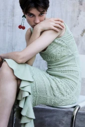 Model Tanya Churbanova wearing a light green knit ruffle dress and beaded cherry earrings, seated against a textured studio wall.