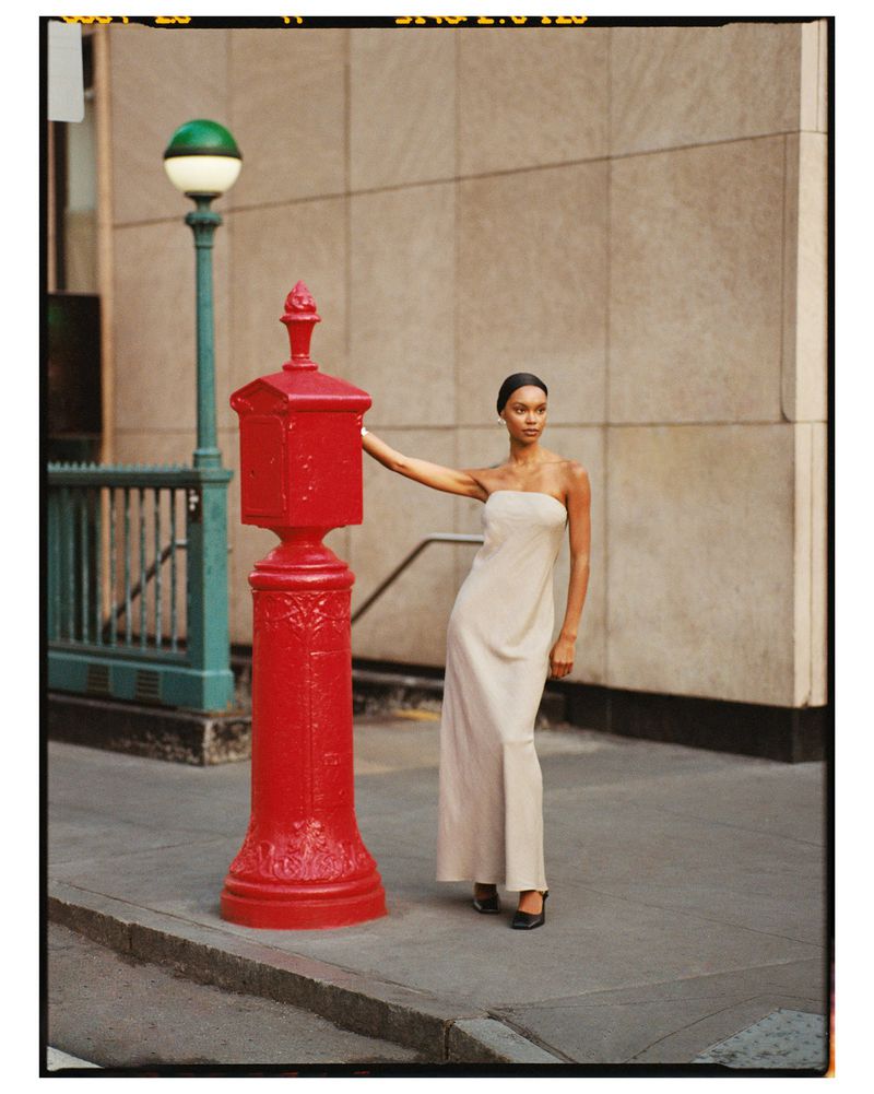 Woman in strapless dress poses next to a vintage red postal box on a city sidewalk with a beige building and green lamp post.