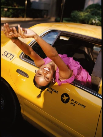 Smiling woman in pink dress leans out of a yellow taxi window in city setting, arms raised, looking toward camera, daytime.