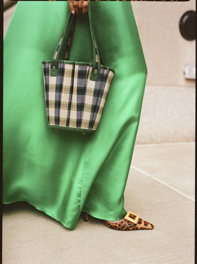 A woman in a vibrant green satin dress showcases a plaid tote bag and leopard-print heels, set against a neutral indoor backdrop.