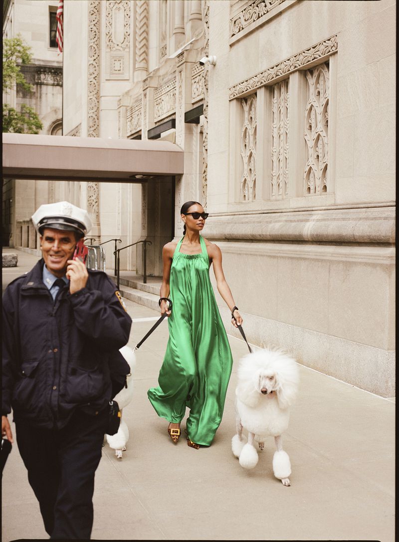 Fashionable woman in a green dress walks two white standard poodles on a city street as a police officer talks on a cell phone.