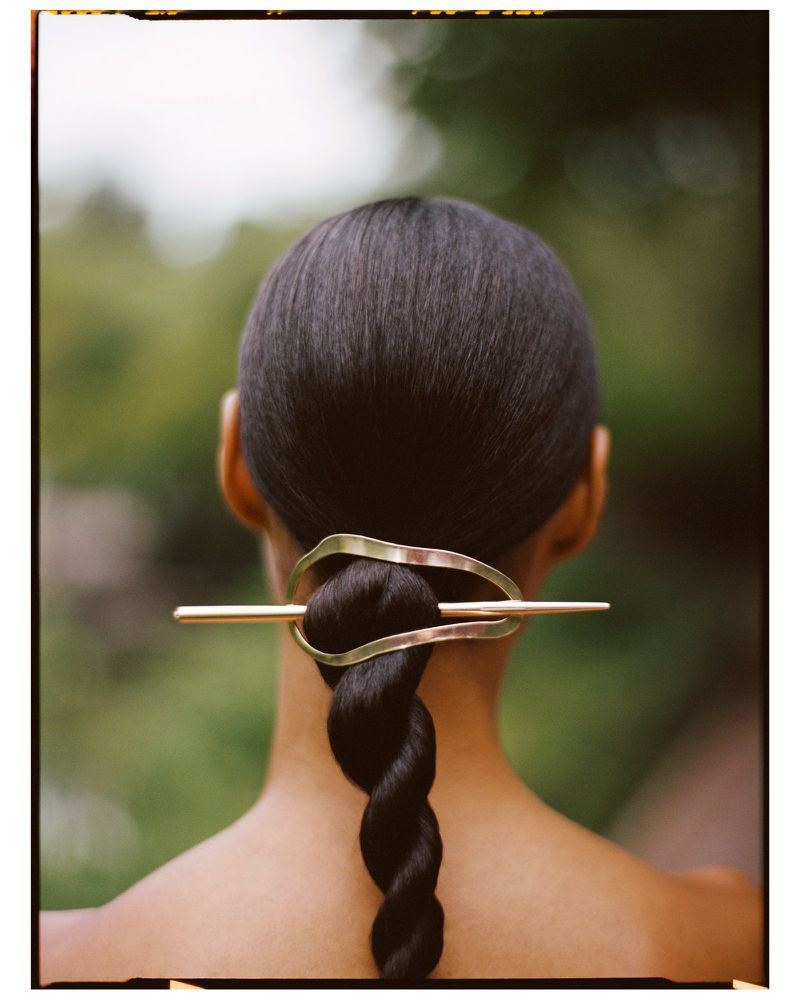 Close-up from the back of an adult female with a dark braided hairstyle accessorized with a gold hair pin, set against a blurred green backdrop.