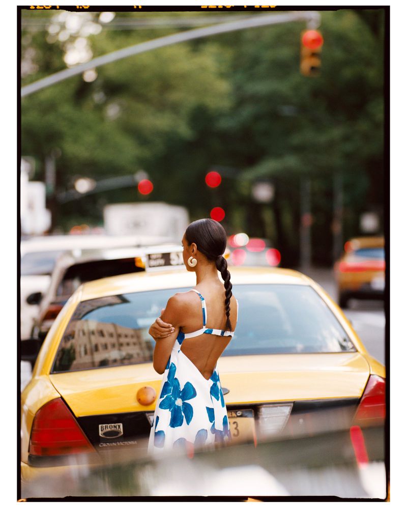 Woman in floral dress stands on a city street with yellow taxicabs and a traffic light in the background on a sunny day.