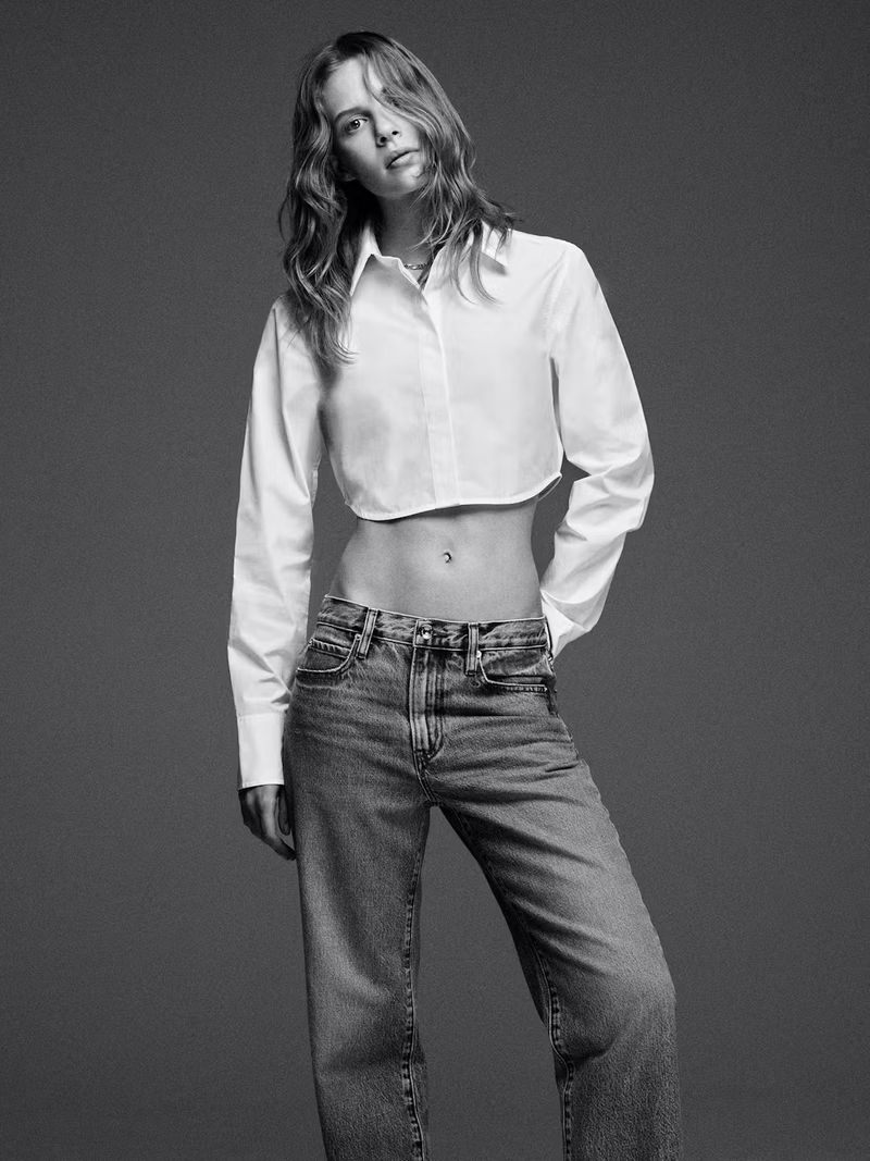 A woman in a white crop top and blue jeans poses in a studio, looking at the camera with a neutral expression in black and white.