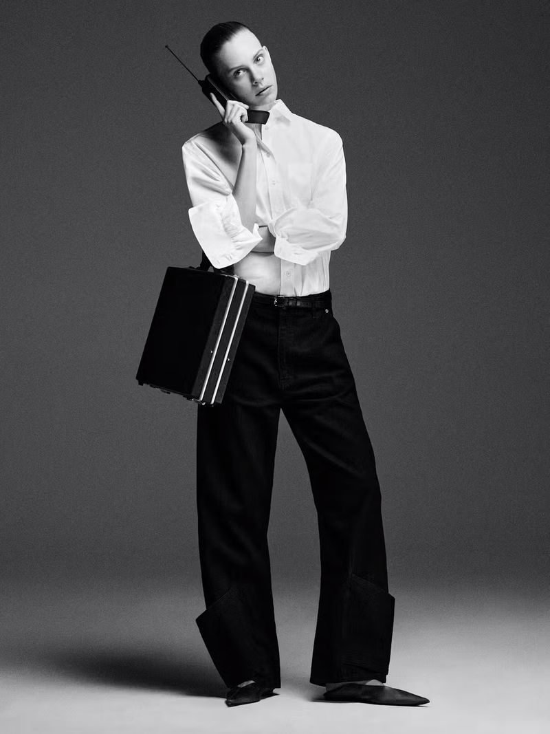 Monochrome fashion portrait of a slender person in a white shirt, black pants and shoes, holding a briefcase and talking on a vintage cell phone.