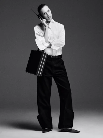 Monochrome fashion portrait of a slender person in a white shirt, black pants and shoes, holding a briefcase and talking on a vintage cell phone.