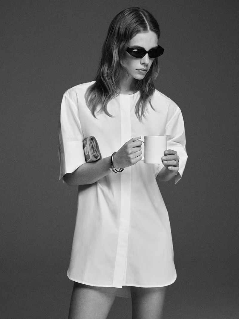 Monochrome fashion shot of a woman with wavy hair wearing sunglasses and a white shirt dress holding a mug and a purse.