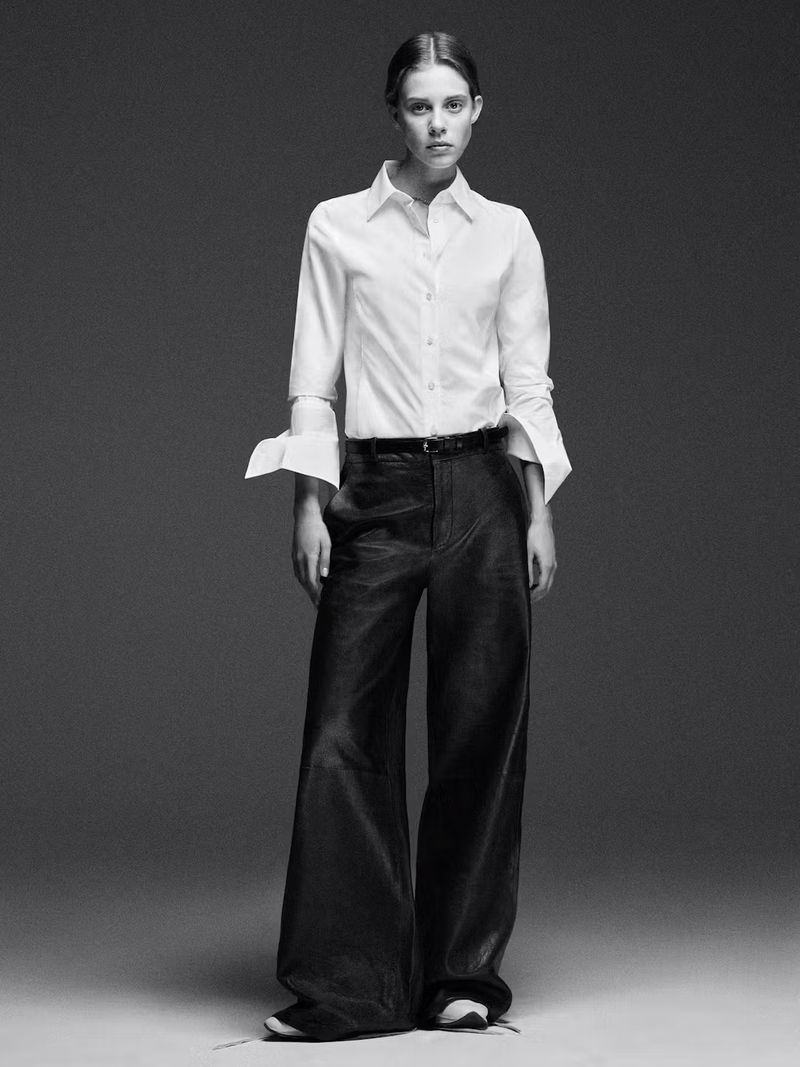 Monochrome portrait of a model wearing a white button-down shirt and dark wide-leg trousers against a grey backdrop
