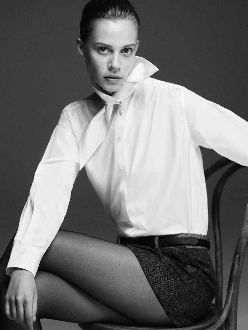 Black and white portrait of a fair-skinned young woman in a white collared shirt and dark shorts, sitting on a chair in a studio setting.