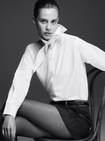 Black and white portrait of a fair-skinned young woman in a white collared shirt and dark shorts, sitting on a chair in a studio setting.