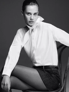 Model wearing a white button-down shirt and dark skirt seated on a wooden chair against a plain studio background.
