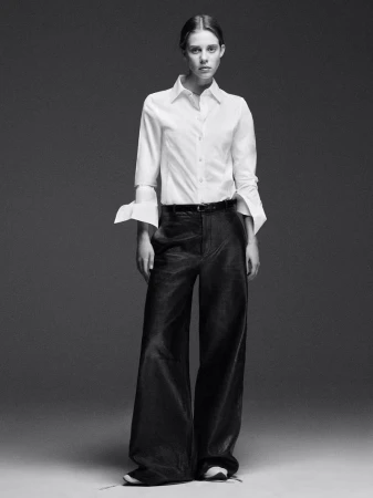 Monochrome portrait of a model wearing a white button-down shirt and dark wide-leg trousers against a grey backdrop