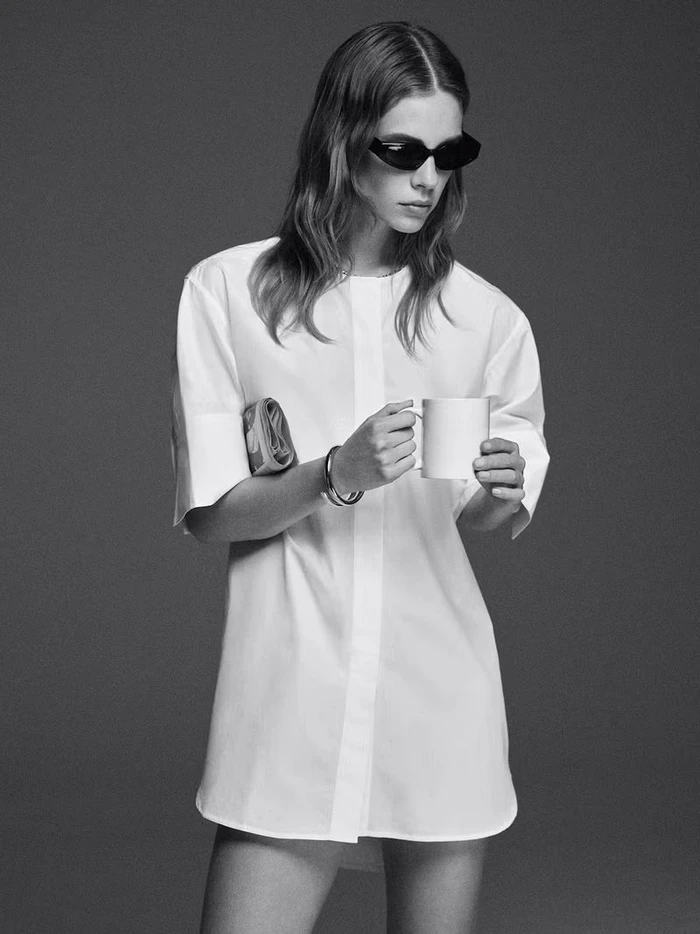 Model in dark sunglasses holding a white mug wearing a short white shirting dress against a grey studio backdrop.