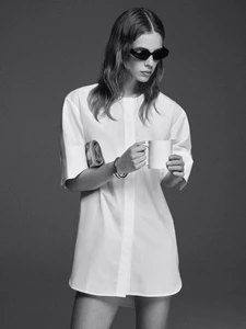 Model in dark sunglasses holding a white mug wearing a short white shirting dress against a grey studio backdrop.