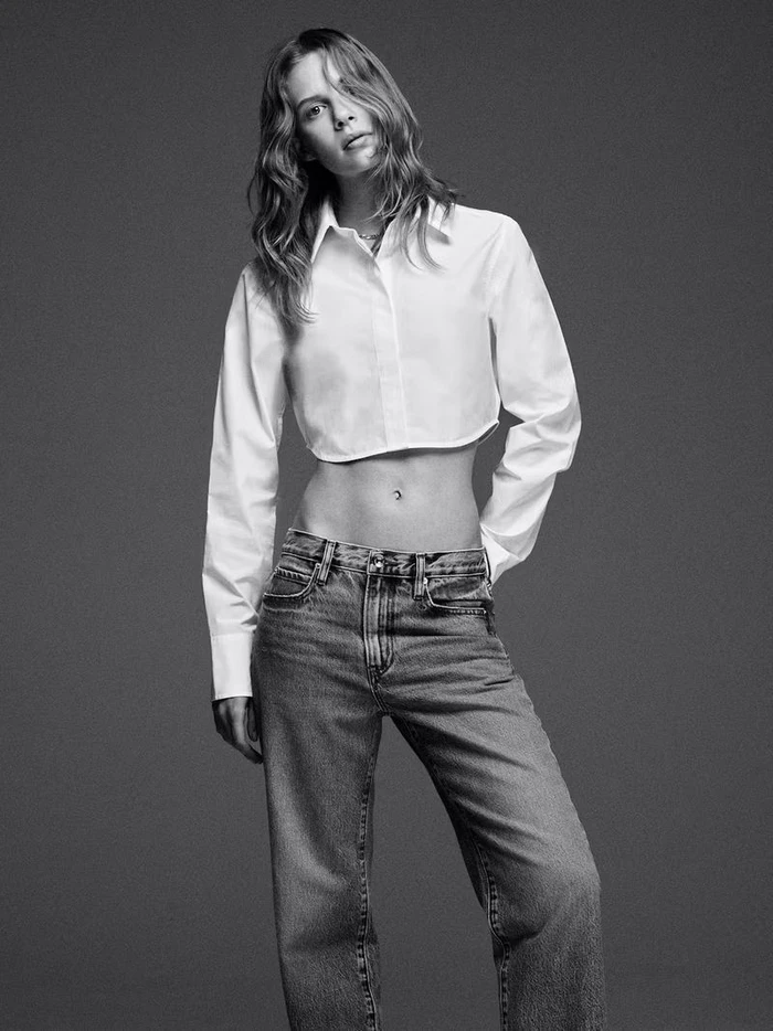 Model wearing a white cropped collared shirt and denim jeans against a grey studio backdrop