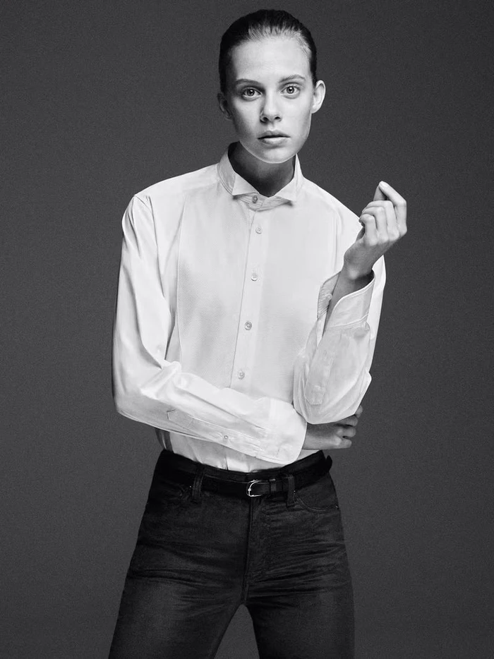Model in white buttoned shirt and dark trousers standing against a seamless dark studio background