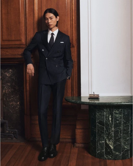 Portrait of a stylish Asian man in a navy pinstripe double-breasted suit, white shirt, and dark tie, leaning against a wooden wall indoors.
