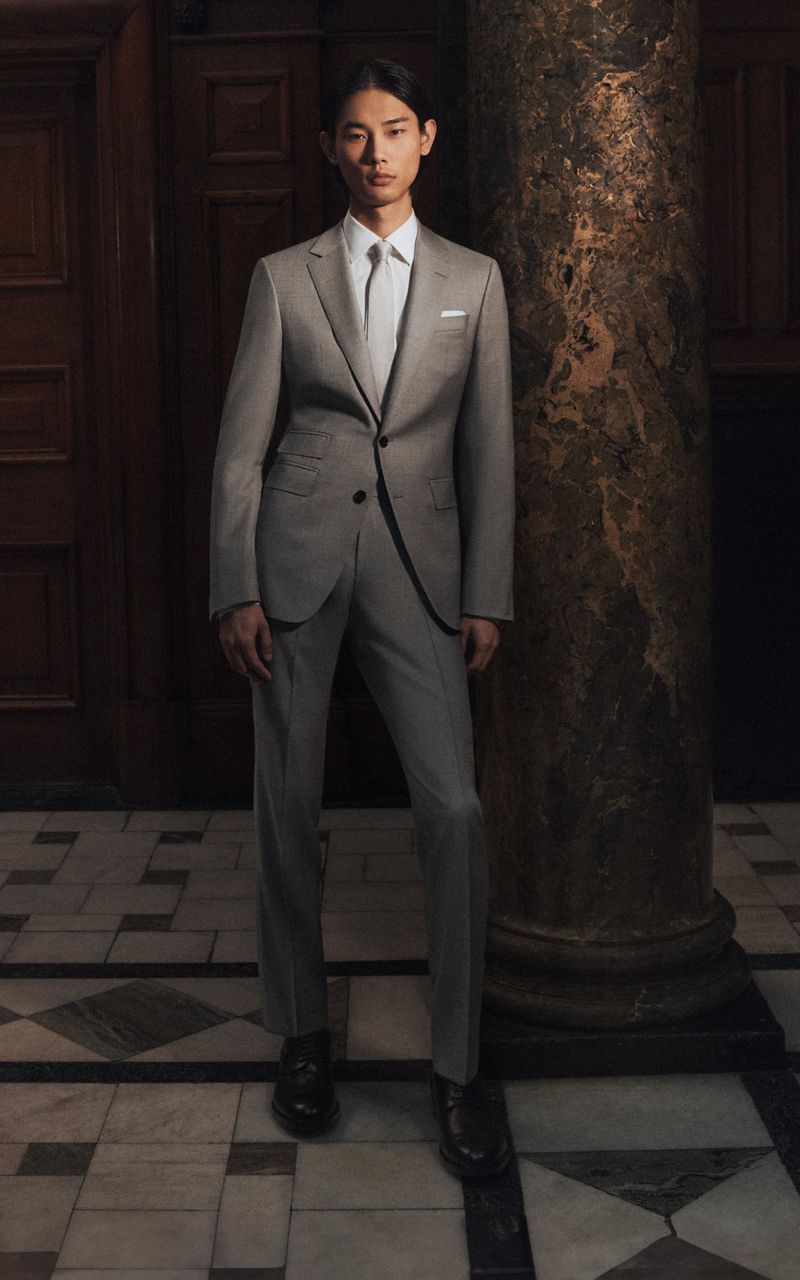 A full shot of a young adult male fashion model in a light grey suit, white shirt, and dark shoes, posing indoors next to a decorative column.