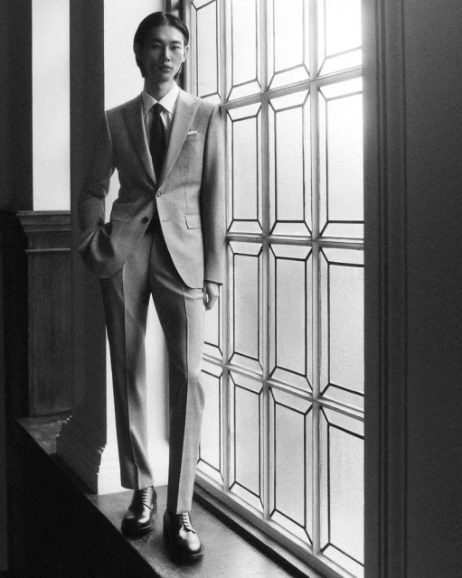 Black and white portrait of an Asian man in a suit posing in front of a window with patterned glass in an indoor setting.