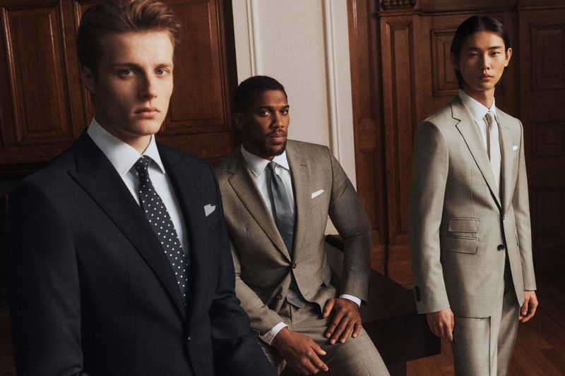 Three men in tailored suits pose indoors, showcasing fashion and style in a softly lit environment with classic wood paneling backdrop.