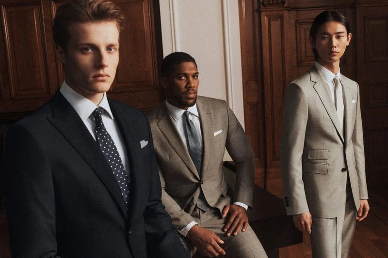 Three men in tailored suits pose indoors, showcasing fashion and style in a softly lit environment with classic wood paneling backdrop.