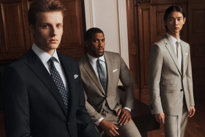 Three men in tailored suits pose indoors, showcasing fashion and style in a softly lit environment with classic wood paneling backdrop.