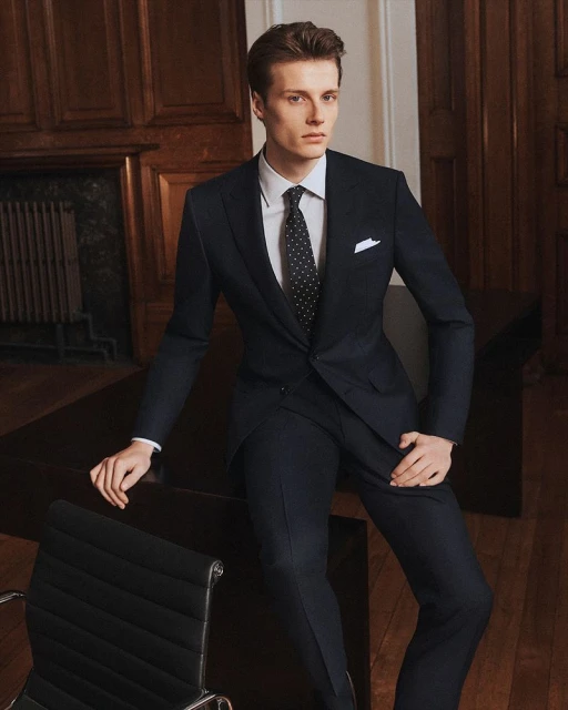 Elegant man in dark suit, white shirt, and patterned tie sitting on desk in office setting, exuding sophistication and professional style.