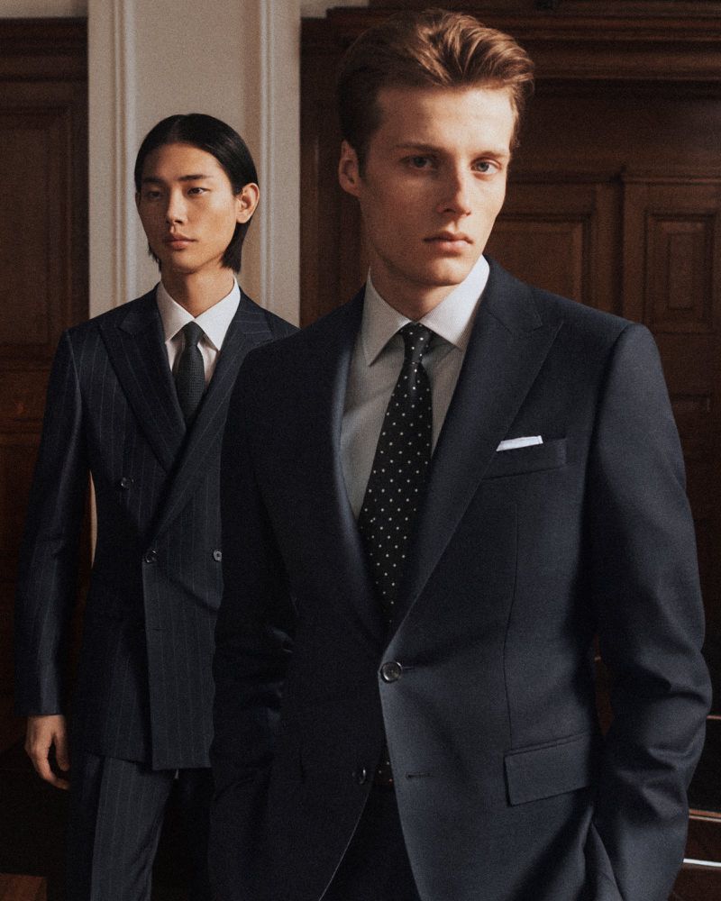 Two men in suits stand indoors. The first man has brown hair, and the second man has black hair and is wearing a pinstripe suit.