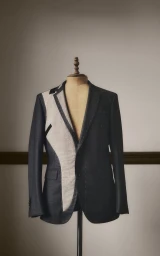 Deconstructed bespoke suit jacket with exposed canvas on a vintage tailor's mannequin against a neutral wall.