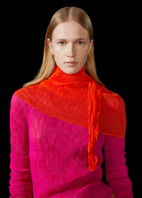Close-up portrait of a fair-skinned adult female in a textured pink and orange long-sleeve top against a dark background.