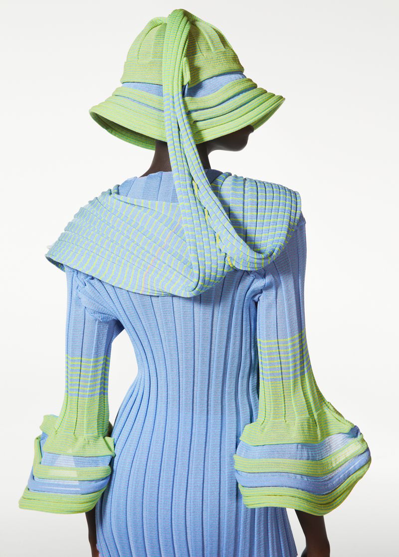Back view of a person wearing a light blue ribbed dress with light green accents and a light green knitted hat in a white background.