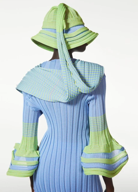 Back view of a person wearing a light blue ribbed dress with light green accents and a light green knitted hat in a white background.