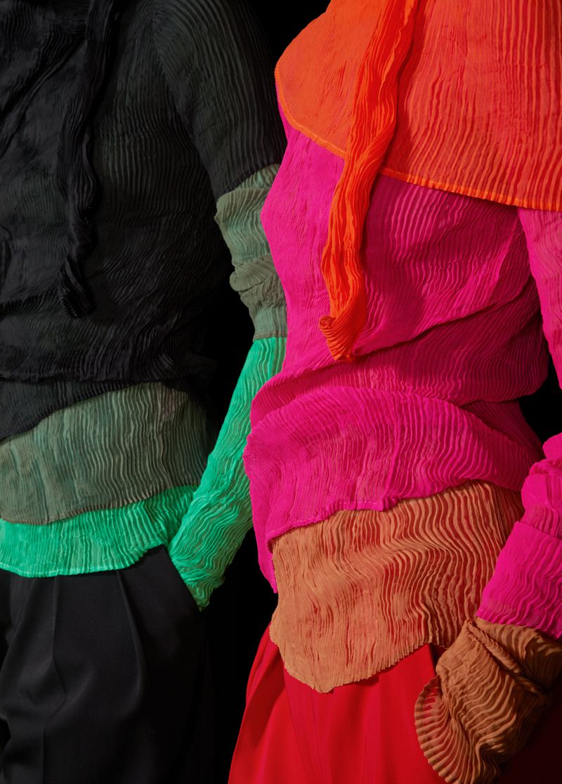 Close-up of two models in colorful pleated tops, vibrant shades of pink, orange, green, and black against a stark black background.