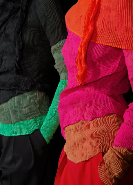 Close-up of two models in colorful pleated tops, vibrant shades of pink, orange, green, and black against a stark black background.
