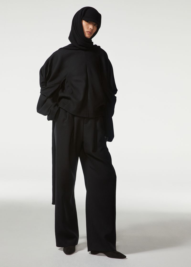 Fashion shot of a person in a black hoodie and wide leg pants with a hooded head covering, standing in a white studio setting.