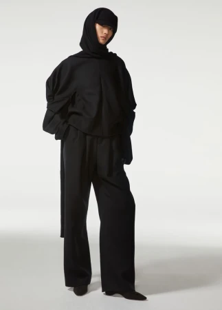Fashion shot of a person in a black hoodie and wide leg pants with a hooded head covering, standing in a white studio setting.