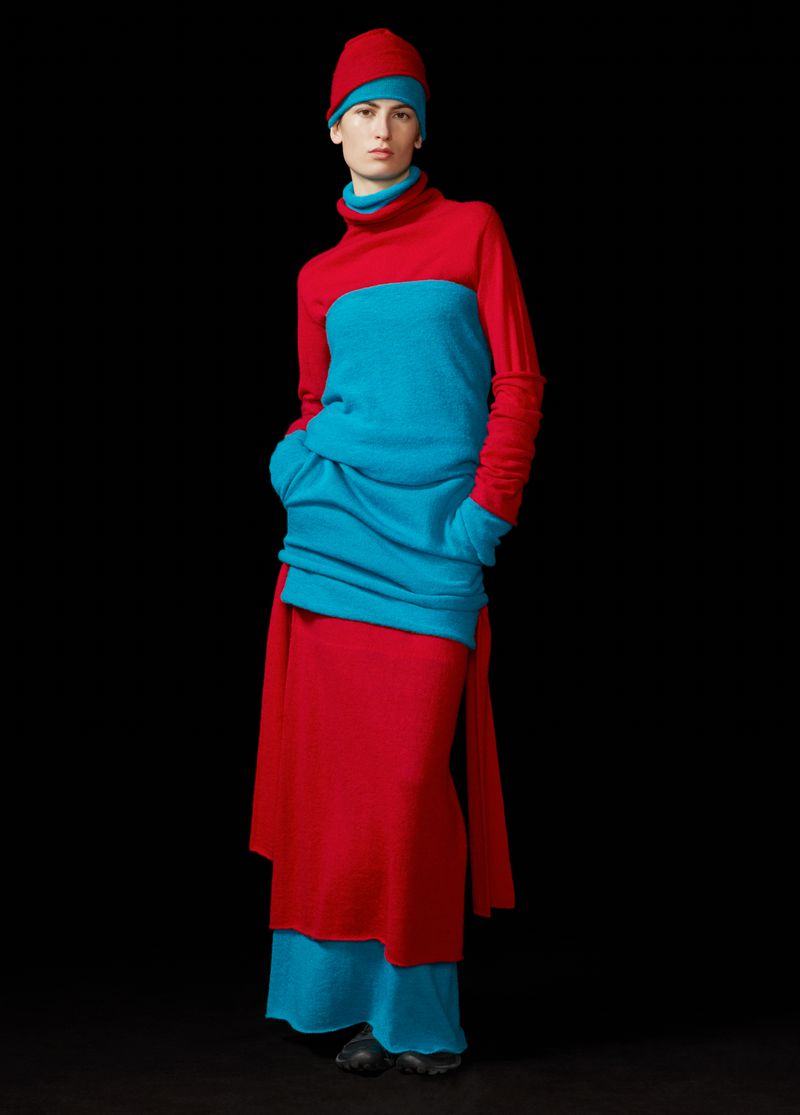 Fashion model poses in a studio wearing a red beanie, red turtleneck top with turquoise panel, red skirt and turquoise under-skirt.