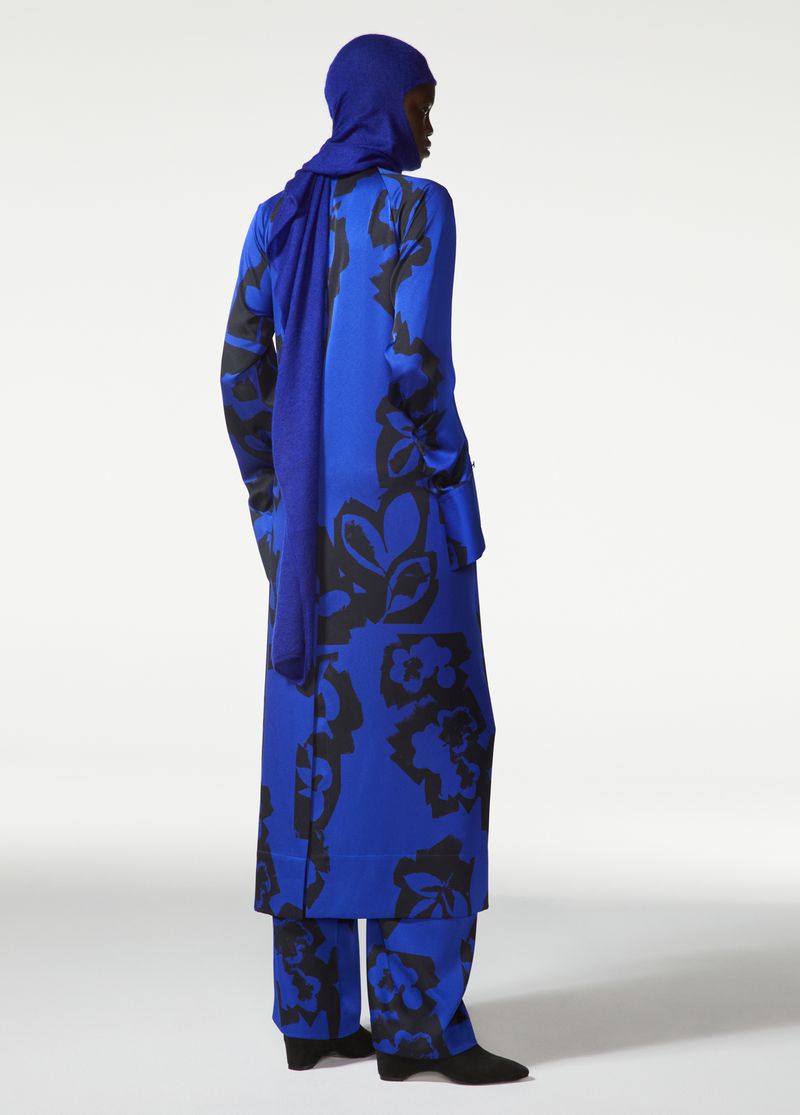 A slender figure models a blue floral print coat and pants with a matching blue head covering against a simple white backdrop in a studio.