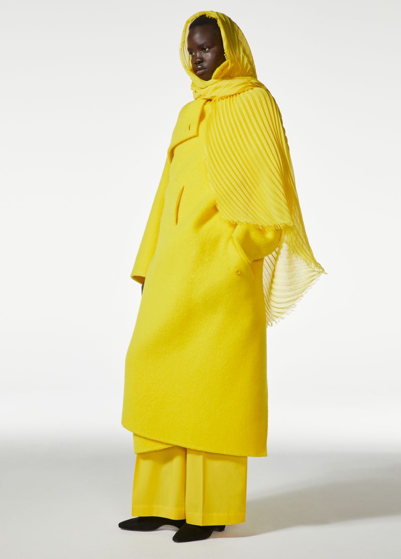 Fashion shot of a woman wearing a yellow coat, scarf, and pants against a white background in a studio setting, full shot.